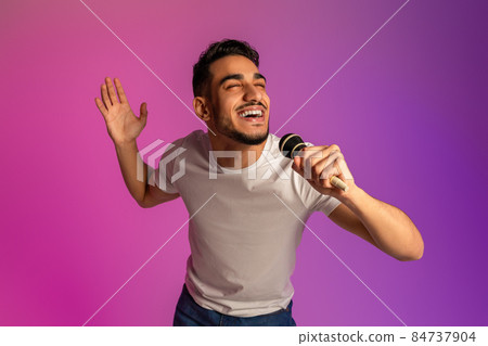Portrait of cool young Arab man singing song, using microphone, performing karaoke in neon light 84737904