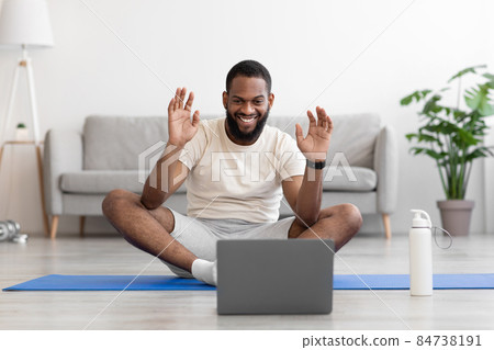 Smiling young black male sit on floor on mat in living room interior with bottle, looks at pc Smiling young black male sit on floor on mat in living room interior with bottle, looks at pc 84738191