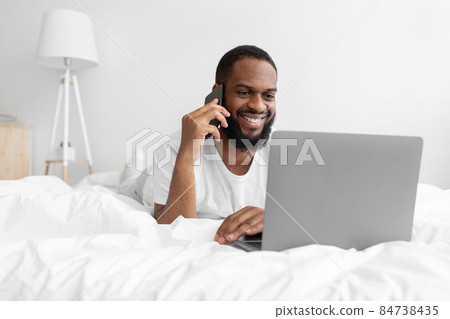 Smiling millennial black bearded guy lies on white bed in bedroom interior speaks by smartphone, looks at laptop Smiling millennial black bearded guy lies on white bed in bedroom interior speaks by smartphone, looks at laptop 84738435