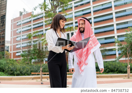 Low angle of businesswoman in formal wear smiling and demonstrating folder with documents to bearded Arab man in traditional clothes while walking on street together 84738732