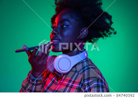 Studio shot of young pretty african girl, student using phone isolated on dark green studio background in purple neon light. Studio shot of young pretty african girl, student using phone isolated on dark green studio background in purple neon light. 84740392