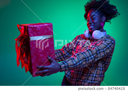 Happy young girl holding big red gift box isolated on dark green studio background in purple neon light. 84740428