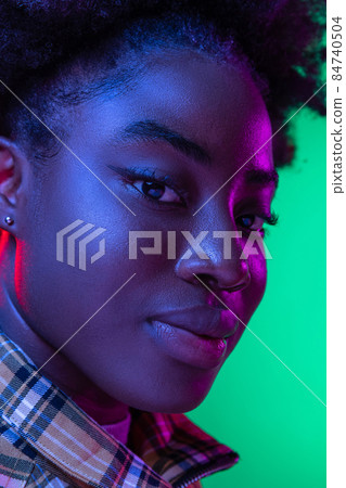 Close-up face of dark skinned young girl looking at camera isolated on dark green studio background in purple neon light. 84740504