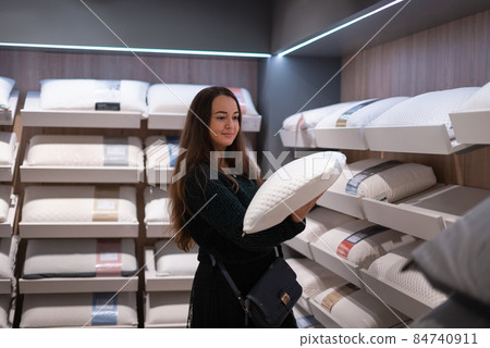 Positive female with long hair inspecting soft pillow while standing near shelves in modern mall 84740911