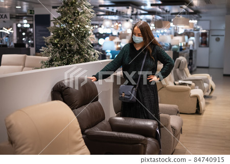 Female customer in mask holding hand on waist and inspecting leather armchair while buying furniture in mall Female customer in mask holding hand on waist and inspecting leather armchair while buying furniture in mall 84740915