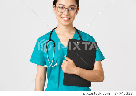 Covid-19, coronavirus disease, healthcare workers concept. Smiling confident asian female nurse, physician in glasses and scrubs holding clipboard, making examination, white background Covid-19, coronavirus disease, healthcare workers concept. Smiling confident asian female nurse, physician in glasses and scrubs holding clipboard, making examination, white background 84743338