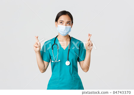 Covid-19, coronavirus disease, healthcare workers concept. Hopeful serious-looking asian doctor, female physician in medical mask and scrubs cross fingers, looking upper left corner Covid-19, coronavirus disease, healthcare workers concept. Hopeful serious-looking asian doctor, female physician in medical mask and scrubs cross fingers, looking upper left corner 84743370