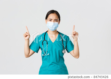 Covid-19, coronavirus disease, healthcare workers concept. Gloomy disappointed asian nurse, physician in medical mask and scrubs, pointing fingers up and frowning perplexed, look frustrated Covid-19, coronavirus disease, healthcare workers concept. Gloomy disappointed asian nurse, physician in medical mask and scrubs, pointing fingers up and frowning perplexed, look frustrated 84743426