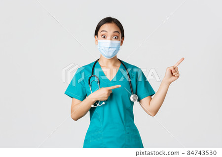 Covid-19, coronavirus disease, healthcare workers concept. Enthusiastic happy asian female doctor, physician or nurse in medical mask and scrubs, pointing upper right corner, show awesome promo 84743530