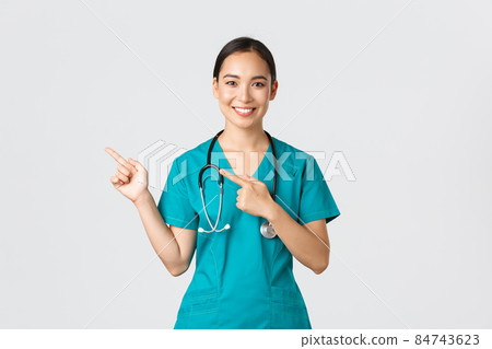 Covid-19, healthcare workers, pandemic concept. Smiling pretty asian doctor, nurse in scrubs and stethoscope, pointing fingers upper left corner, showing online clinic banner, white background Covid-19, healthcare workers, pandemic concept. Smiling pretty asian doctor, nurse in scrubs and stethoscope, pointing fingers upper left corner, showing online clinic banner, white background 84743623