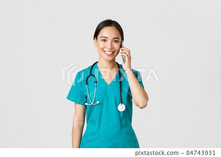 Covid-19, healthcare workers and preventing virus concept. Smiling happy asian female doctor, young intern in scrubs talking on phone, looking at camera hopeful, white background 84743931