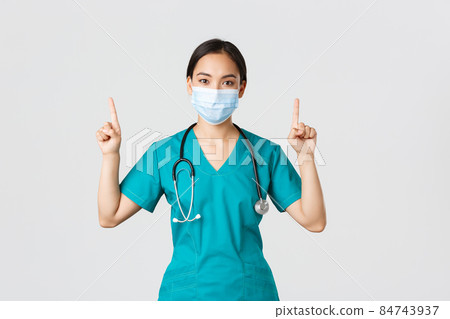 Covid-19, coronavirus disease, healthcare workers concept. Young professional asian female doctor, nurse in medical mask and scrubs, pointing fingers up, showing way, advertising Covid-19, coronavirus disease, healthcare workers concept. Young professional asian female doctor, nurse in medical mask and scrubs, pointing fingers up, showing way, advertising 84743937