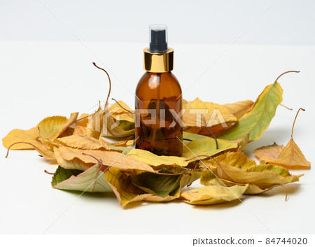 glass brown bottle with cosmetic spray on white background in the middle of yellow leaves 84744020