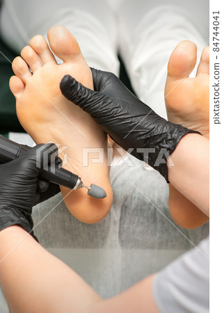 Peeling female feet. Hands of podiatry specialist in black gloves using special grinding equipment polish female foot from callus and corn in the beauty salon. 84744041
