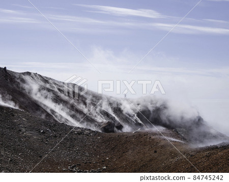Silhouette of man hiking at coldera of Avachinsky stratovolcano, also known as Avacha Volcano. Backpacker tourist moves upon rocks behind steam from hot geysers. Kamchatka Peninsula, Russia. Silhouette of man hiking at coldera of Avachinsky stratovolcano, also known as Avacha Volcano. Backpacker tourist moves upon rocks behind steam from hot geysers. Kamchatka Peninsula, Russia. 84745242