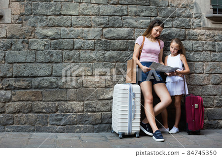 Woman and little girl traveler with suitcase leaning against stone wall Woman and little girl traveler with suitcase leaning against stone wall 84745350
