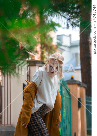 Fashionable beautiful young woman with blond hair in a stylish long coat, checkered pants, red shoes and glasses poses in the city streets. 84745744
