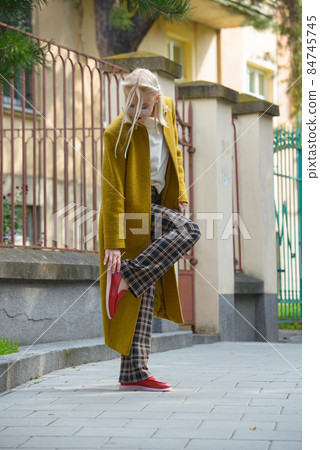 Fashionable beautiful young woman with blond hair in a stylish long coat, checkered pants, red shoes and glasses poses in the city streets. 84745745