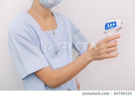 Temperature measurement gun in doctor hands. Close-up shot of doctor wearing protective surgical mask ready to use infrared isometric thermometer gun to check body temperature for virus symptoms. Temperature measurement gun in doctor hands. Close-up shot of doctor wearing protective surgical mask ready to use infrared isometric thermometer gun to check body temperature for virus symptoms. 84746320