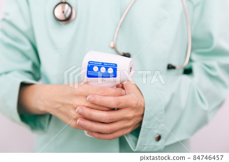 Temperature measurement gun in doctor hands. Close-up shot of doctor wearing protective surgical mask ready to use infrared isometric thermometer gun to check body temperature for virus symptoms. 84746457