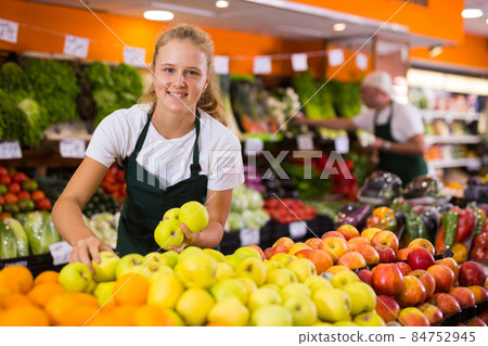 Fifteen-year-old girl who works part-time as a trainee saleswoman puts apples on the counter 84752945
