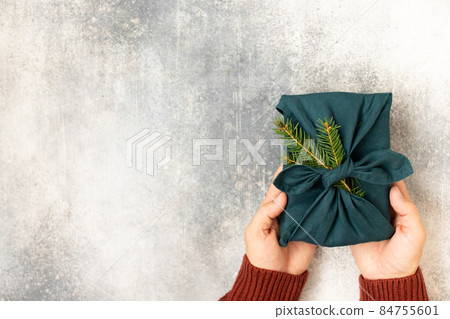 Furoshiki-style gift and spruce for Christmas or New Year on a grey background with hands. Flat lay. Copy space. Furoshiki-style gift and spruce for Christmas or New Year on a grey background with hands. Flat lay. Copy space. 84755601