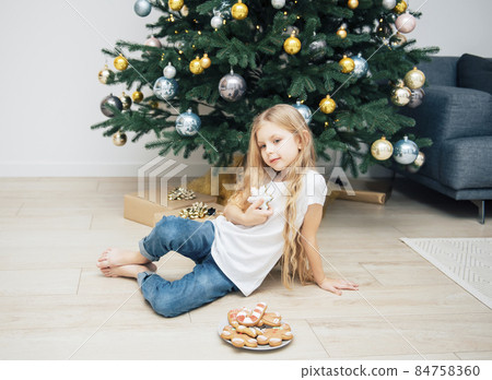 Girl eating Christmas gingerbread near the Christmas tree Girl eating Christmas gingerbread near the Christmas tree 84758360