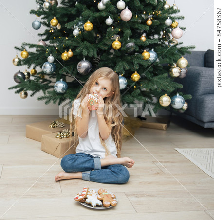 Girl eating Christmas gingerbread near the Christmas tree Girl eating Christmas gingerbread near the Christmas tree 84758362