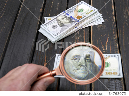 One hundred dollar bill being studied through a magnifying glass on a old black wooden table One hundred dollar bill being studied through a magnifying glass on a old black wooden table 84762075