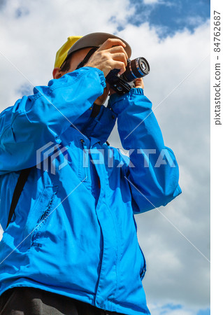 A man in a blue jacket and yellow cap with a camera in his face. A photographer against a cloudy sky. A travel blogger with a camera. View from below A man in a blue jacket and yellow cap with a camera in his face. A photographer against a cloudy sky. A travel blogger with a camera. View from below 84762687