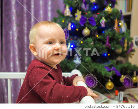 A little cheerful baby girl at a Christmas tree. A little cheerful baby girl at a Christmas tree. 84763116