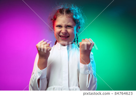 Portrait of angry little girl, kid clenches fists isolated on gradient purple blue color background in neon light Portrait of angry little girl, kid clenches fists isolated on gradient purple blue color background in neon light 84764623