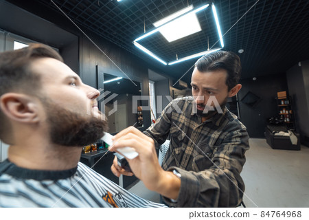 Barber works with a beard clipper. Hipster client getting haircut. 84764968