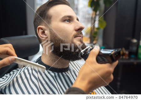 Barber works with a beard clipper. Hipster client getting haircut. Barber works with a beard clipper. Hipster client getting haircut. 84764970