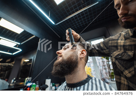 Barber works with a beard clipper. Hipster client getting haircut. 84764976