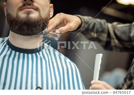 Hairdresser doing haircut of beard using comb and scissors Hairdresser doing haircut of beard using comb and scissors 84765017
