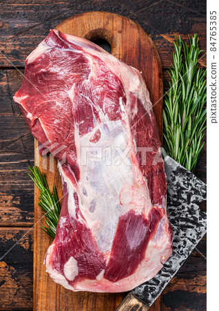 Raw lamb or goat shoulder meat on the bone on a wooden butcher cutting board with cleaver. Dark wooden background. Top view Raw lamb or goat shoulder meat on the bone on a wooden butcher cutting board with cleaver. Dark wooden background. Top view 84765385