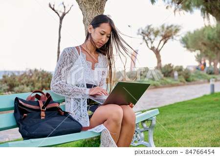 Beautiful young Asian woman using laptop sitting on an outdoor bench 84766454