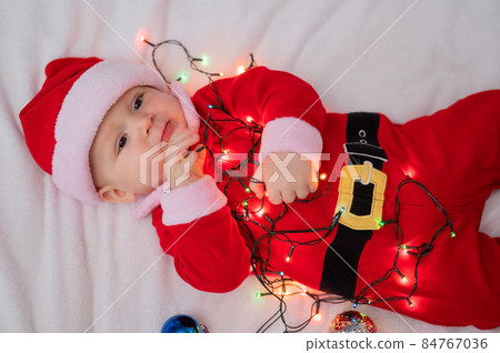A baby boy dressed in a santa suit lies on the bed and plays with a Christmas garland. 84767036
