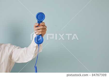 Hand of unknown man holds blue wired retro phone which is symbol of emergency call. Retro telephone handset on light blue background with empty space for text on right. Helpline concept. Banner. 84767309