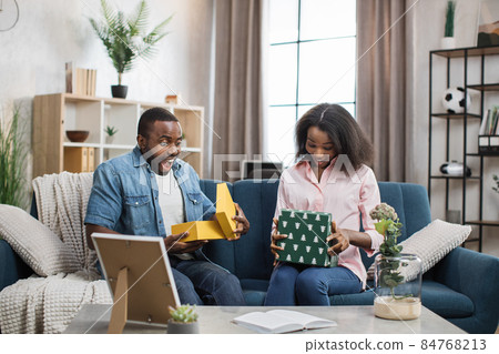 Surprised couple opening gift boxes while sitting on couch Surprised couple opening gift boxes while sitting on couch 84768213