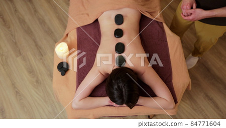 Top view of beautiful young woman gets hot stones massage at spa. Attractive female relaxing at spa center with a stone therapy. 84771604