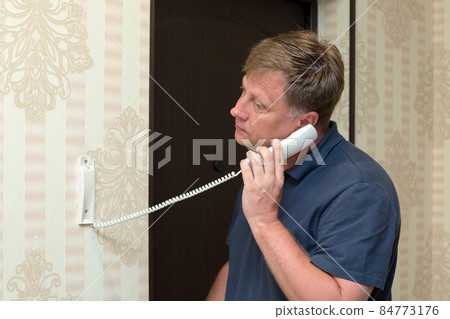 An adult blond man is talking on the intercom in the hallway of his apartment 84773176