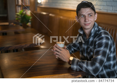 Smiling young caucasian man in checkered shirt and cap drinks coffee in a bar. Coffee shop, cafe, latte, caramel cappuccino. High quality photo Smiling young caucasian man in checkered shirt and cap drinks coffee in a bar. Coffee shop, cafe, latte, caramel cappuccino. High quality photo 84774734