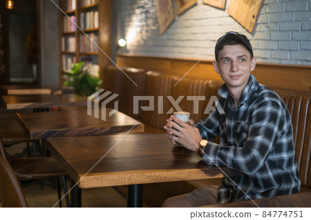 Smiling young caucasian man in checkered shirt and cap drinks coffee in a bar. Coffee shop, cafe, latte, caramel cappuccino. High quality photo Smiling young caucasian man in checkered shirt and cap drinks coffee in a bar. Coffee shop, cafe, latte, caramel cappuccino. High quality photo 84774751