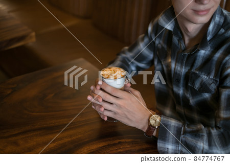 Man in checkered shirt drinks coffee in a bar. Holding caramel latte in his hands. Close up, Coffee shop, cafe, latte, caramel cappuccino. Man in checkered shirt drinks coffee in a bar. Holding caramel latte in his hands. Close up, Coffee shop, cafe, latte, caramel cappuccino. 84774767