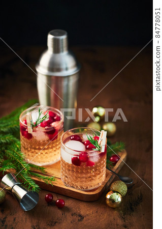 Alcoholic cocktail or non-alcoholic cocktail with vodka and cranberries with ice, shaker, jigger and bar spoon, fir branches and glowing garland for christmas on wooden background Alcoholic cocktail or non-alcoholic cocktail with vodka and cranberries with ice, shaker, jigger and bar spoon, fir branches and glowing garland for christmas on wooden background 84778051