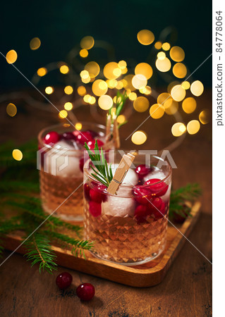 Alcoholic cocktail or non-alcoholic cocktail with vodka and cranberries with ice with fir branches and glowing garland for christmas on wooden background Alcoholic cocktail or non-alcoholic cocktail with vodka and cranberries with ice with fir branches and glowing garland for christmas on wooden background 84778064