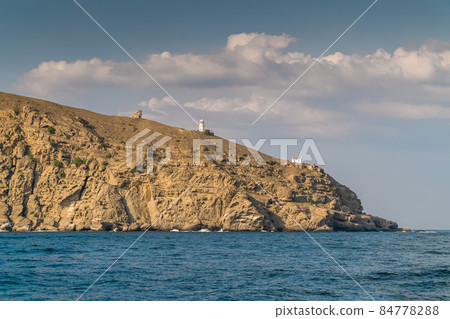 The Republic of Crimea. July 21, 2021. View from the Black Sea in warm sunny weather on the lighthouse and buildings on Mount Meganom, which is located near the city of Sudak. The Republic of Crimea. July 21, 2021. View from the Black Sea in warm sunny weather on the lighthouse and buildings on Mount Meganom, which is located near the city of Sudak. 84778288