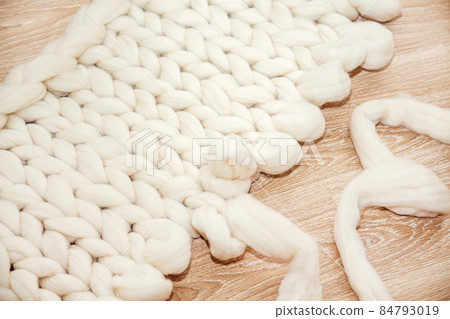 The process of knitting a plaid of large knitting from merino wool. Unfinished work with open loops on wooden floor 84793019